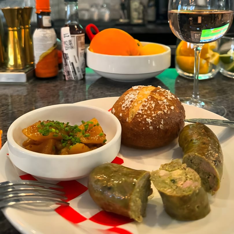 A plate of sliced sausage, a side of potatoes with chives, and a pretzel bun, with a glass of white wine.
