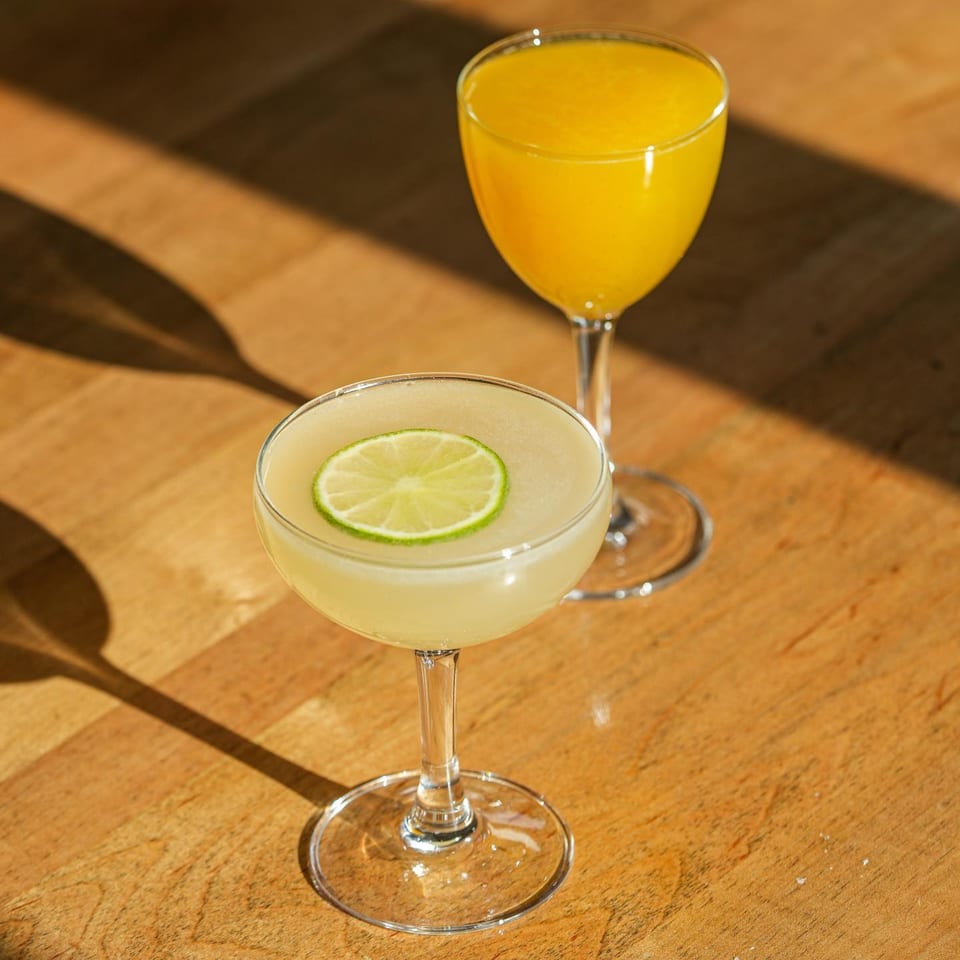 Two cocktails on a wooden table, one yellow with a lime slice, the other orange.