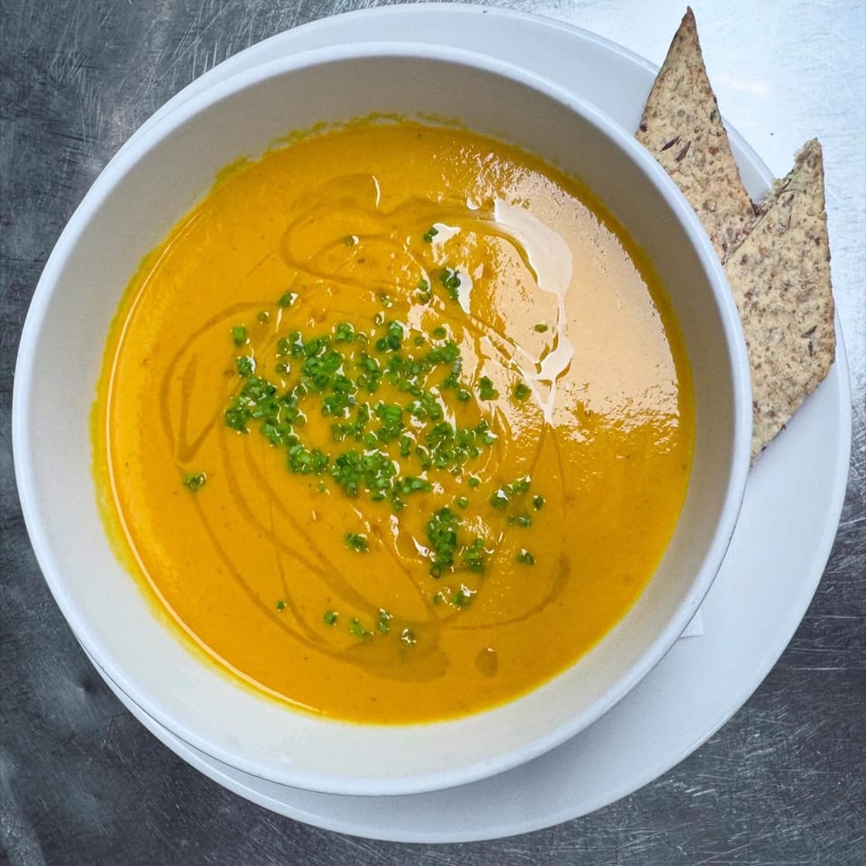 A bowl of vibrant orange soup, garnished with chives and olive oil, served with two crackers.