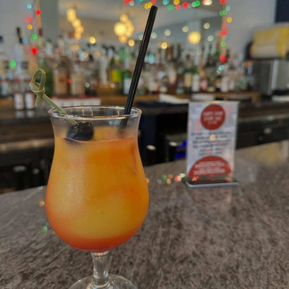 A vibrant orange and red cocktail with a cherry and black straw sits on a bar.