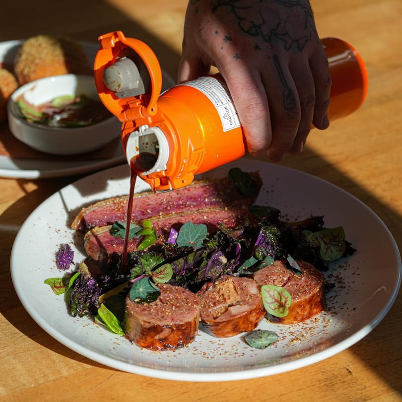 A tattooed hand pours sauce from an orange bottle onto sliced meat and purple kale on a white plate.